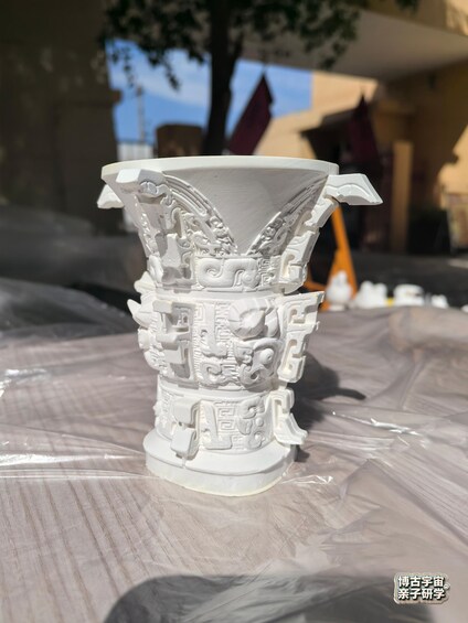 A white intricately carved vase on a table with a plastic sheet underneath. 5