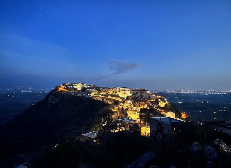 A coastal town at dusk with illuminated buildings and a prominent hilltop structure. 3