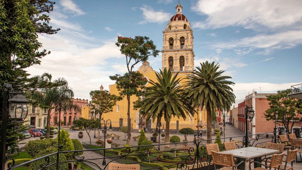 A plaza with a prominent church tower, palm trees, and café seating. 1