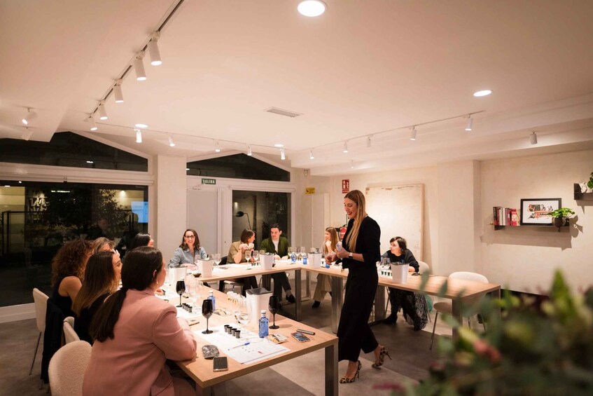 A conference room with people seated at tables, a speaker addressing the group, and a sign reading 'SALIDA' on the wall. 2