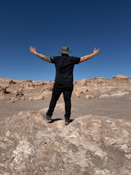 A person standing on rocky terrain with arms outstretched, wearing a hat, black shirt, and jeans. 4