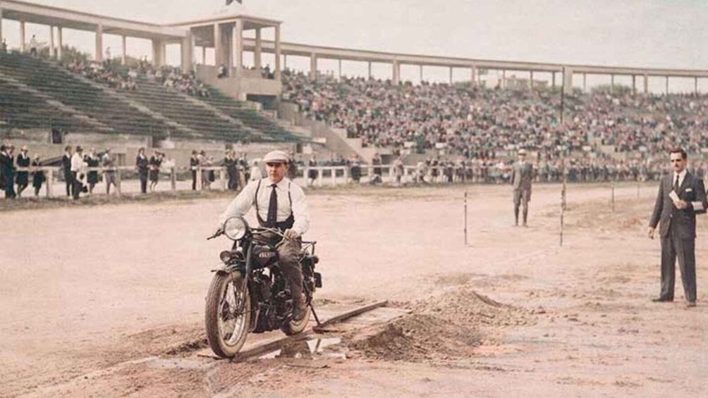 A vintage motorcycle race with a rider on a dirt track, spectators in the stands, and a bridge in the background. 1