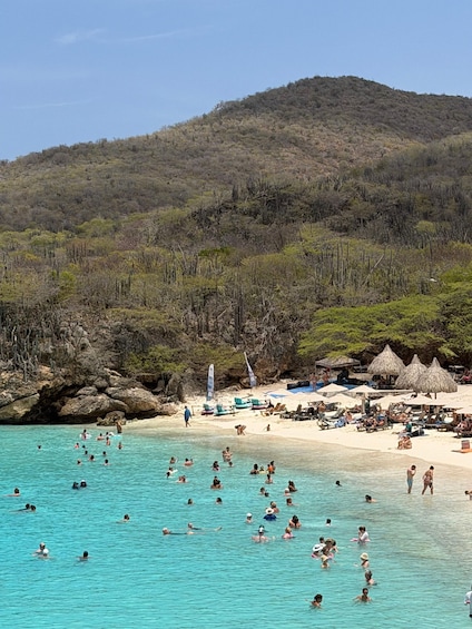 A beach with clear turquoise water, people swimming and sunbathing, and a backdrop of a hilly landscape with sparse vegetation. 5