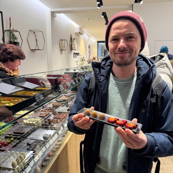 A person holding a tray of assorted pastries in a bakery with various baked goods displayed in the background. 1