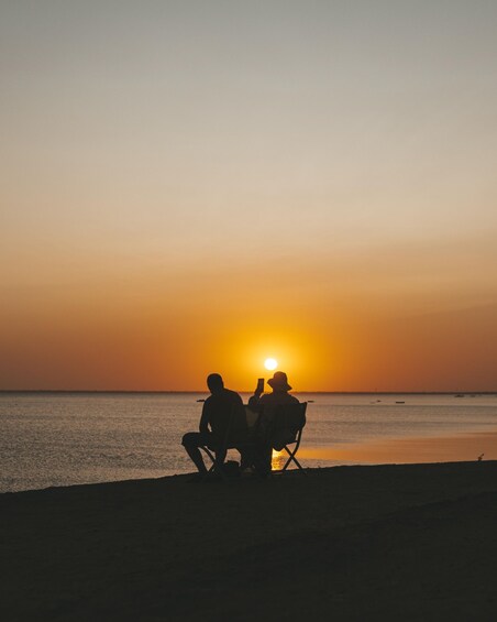 Two people sitting on beach chairs facing the sunset over the ocean. 2