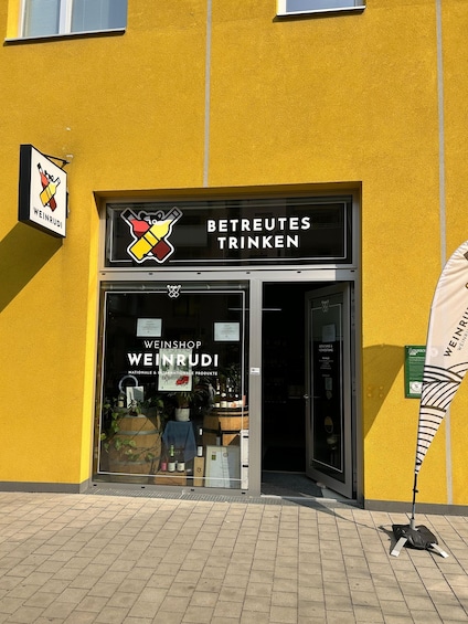 A storefront with a yellow wall and a sign that reads 'BETREUTES TRINKEN' and 'WEINSHOP WEINRUDI.' 2