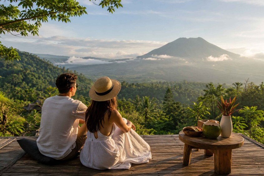 A couple sitting on a wooden deck overlooking a mountainous landscape with a volcano in the distance. 2