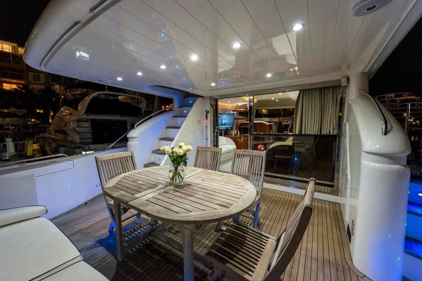 A yacht's deck with a round wooden table, chairs, a vase of flowers, and a glass-enclosed area with a TV. 4
