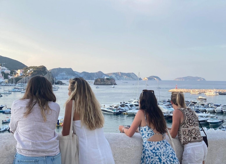 A group of women standing by a marina with boats and rocky hills in the background. 1
