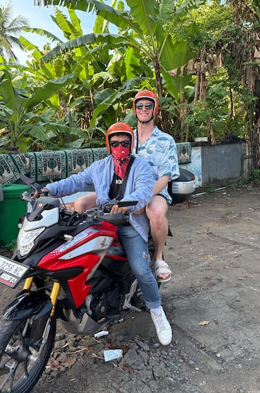 Two people on a red motorcycle with a license plate, surrounded by lush greenery and a concrete wall. 1
