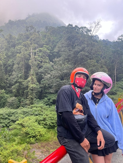 Two people wearing helmets, sitting on a red structure with a forested mountain in the background. 4