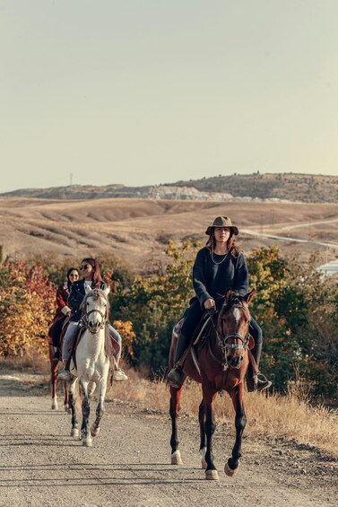 Three people riding horses on a dirt road with autumn foliage and hills in the background. 1