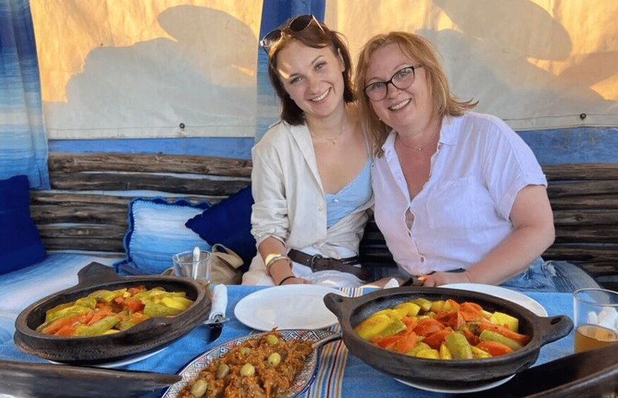 Two people seated at a table with a meal consisting of vegetables and a dish with meat and olives. 5
