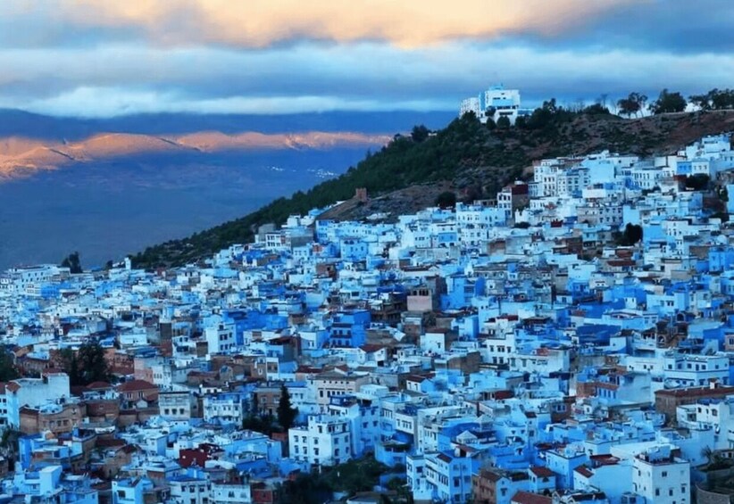 A densely packed town with predominantly white buildings, a prominent hilltop structure, and a mountainous backdrop. 2