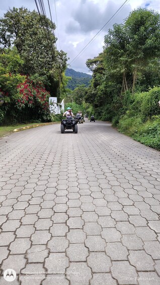 A paved road with ATVs in the distance, surrounded by lush greenery and hills. 1