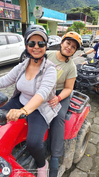 Two people on a red ATV with a building and parked cars in the background. 2