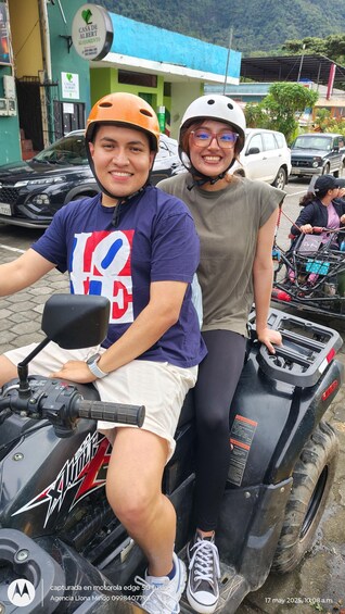Two people on an ATV, with a building and parked cars in the background. 3