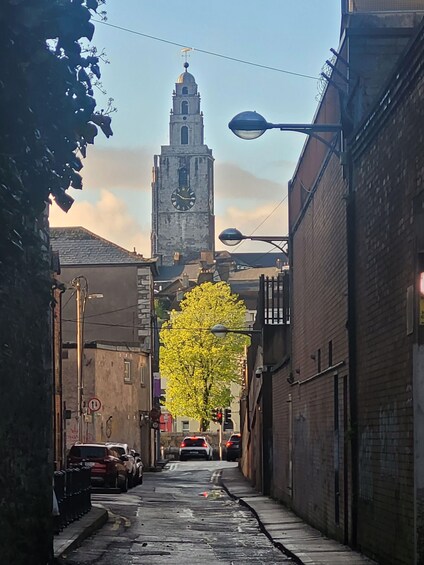 A narrow street with parked cars, a tall clock tower, and a yellow flowering tree. 3