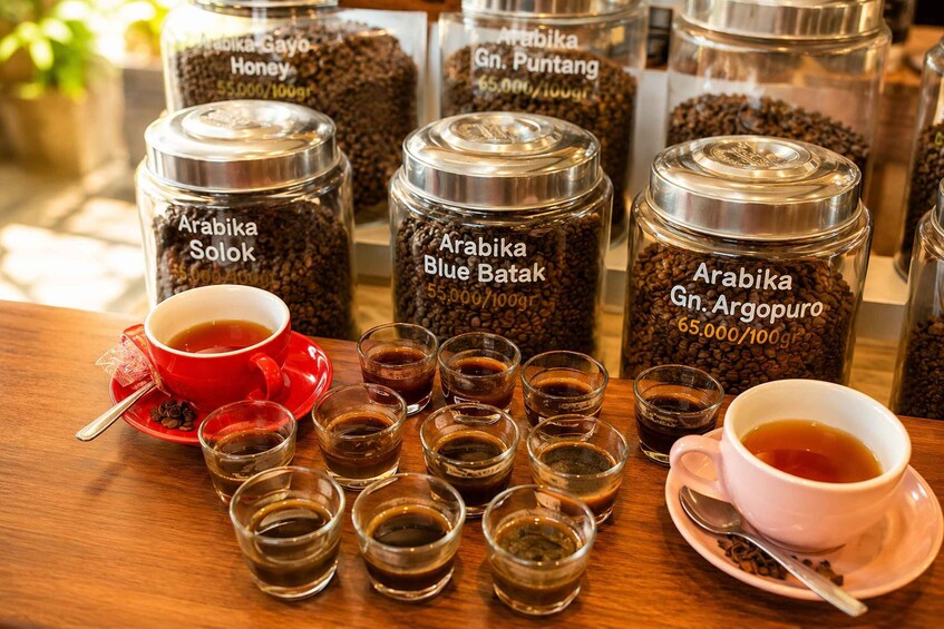 Coffee beans in jars labeled with their names and roast levels, cups of coffee, and a spoon on a table. 3