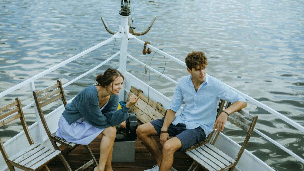 Two people sitting on a wooden boat with metal railings, surrounded by calm water. 4
