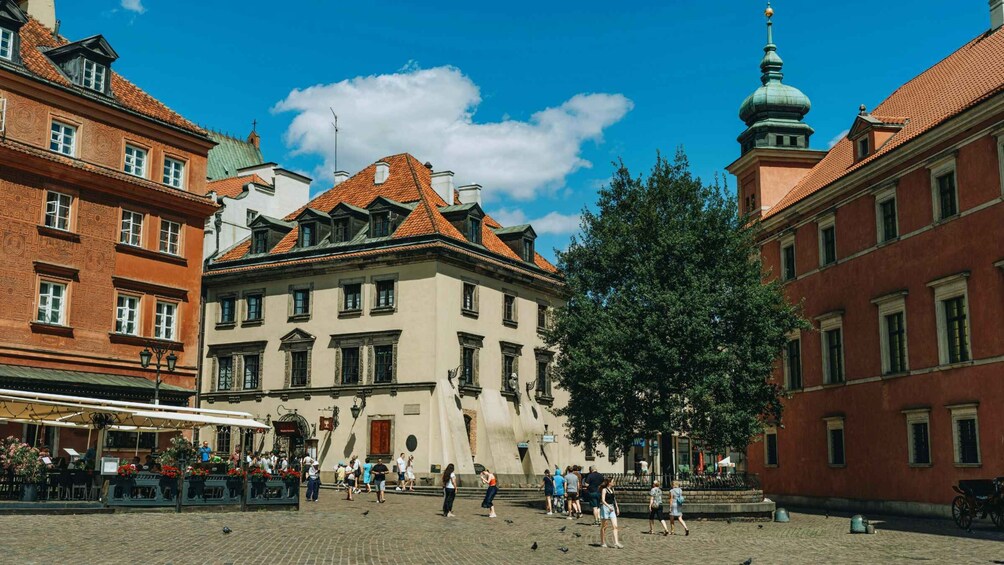 A cobblestone square with historic buildings, a large tree, and people walking. 3