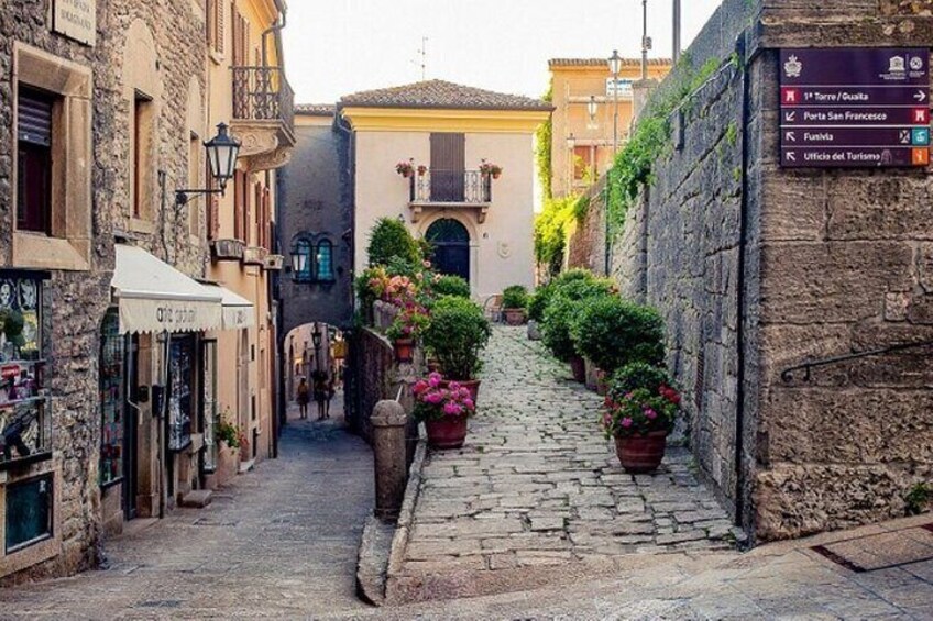 A narrow cobblestone street lined with buildings, potted plants, and a signpost with directions. 3