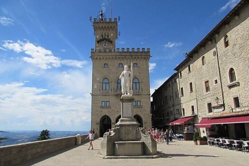 A historic tower with a statue base, a stone wall, and a building with red awnings. 5