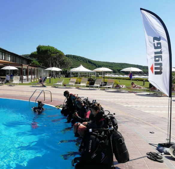 A group of scuba divers preparing by the poolside, with a flag and umbrellas in the background. 1