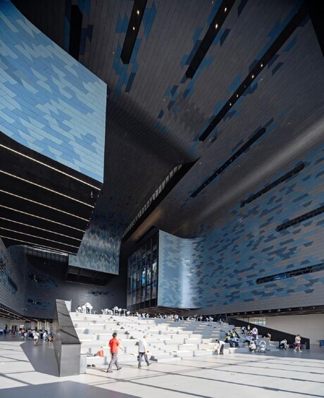 A modern architectural space with a patterned ceiling, a large digital display, and a seating area with people. 3