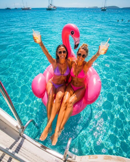 Two people in a pool with a pink flamingo float, surrounded by clear blue water and boats in the background. 3