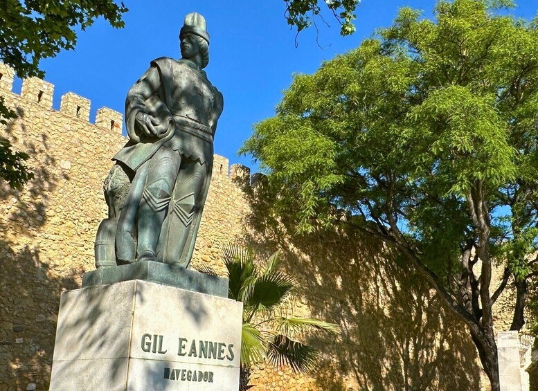 A statue of Gil Eannes, a historical figure, with a stone wall and trees in the background. 2