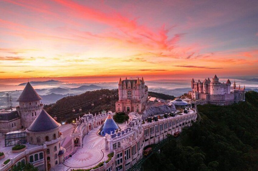 A castle complex with multiple towers and a large central building, surrounded by lush greenery and overlooking a valley. 1