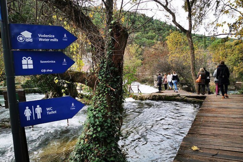 A wooden boardwalk over a stream, with signs pointing to Vodenice Water Mills, Suvenirnica Gift Shop, and WC Toilets. 5