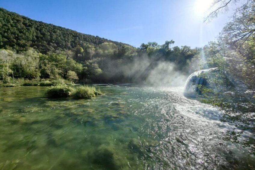 A clear river with a waterfall, surrounded by dense forest and hills. 4