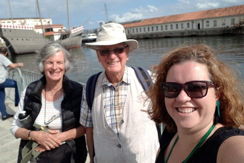 Three people standing by a waterfront with boats and a historic building in the background. 3