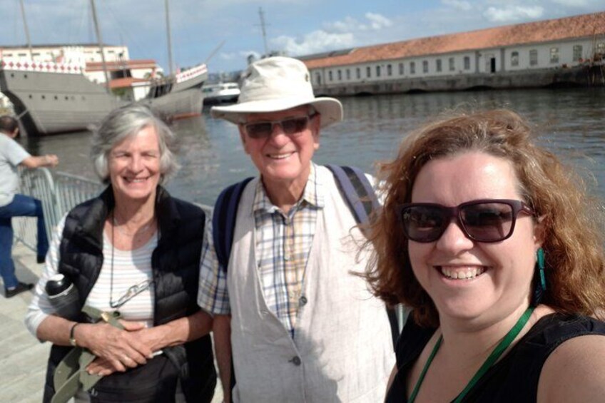 Three people standing by a waterfront with boats and a historic building in the background. 3