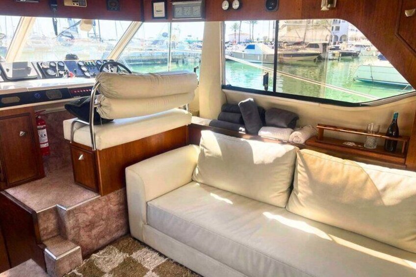 A yacht interior with a white sofa, a steering wheel, and a view of the marina outside. 3