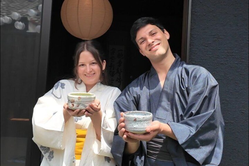 Two people holding ceramic bowls, standing in front of a building with a dark facade and a hanging paper lantern. 1