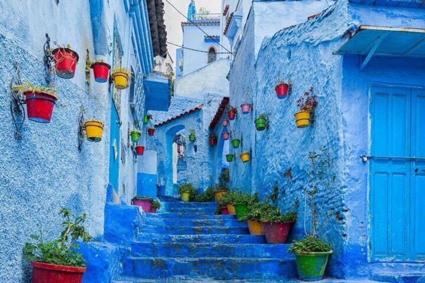 A narrow alley with blue-painted buildings, colorful hanging pots, and a blue door. 5