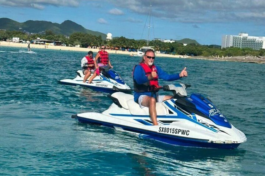 Two people jet-skiing on a body of water with a beach and buildings in the background. 1