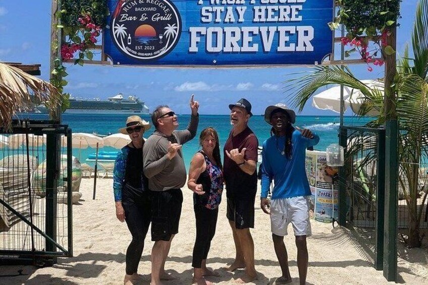 A group of people standing in front of a sign that reads 'Stay Here Forever' at a beachside location. 2