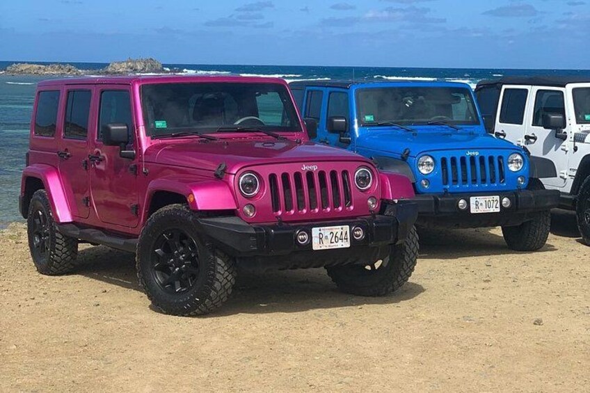 A pink and a blue Jeep parked on a sandy beach with the ocean in the background. 1