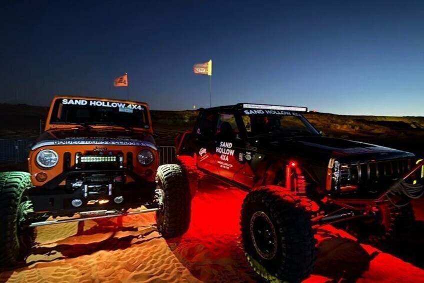 Two off-road vehicles with 'SAND HOLLOW 4X4' written on them, parked on a sandy terrain with a flag in the background. 4