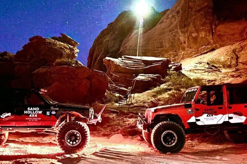 A red off-road vehicle with 'SAND HOLLOW 4x4' written on it, parked in a desert-like environment with rocky terrain and a starry sky. 5