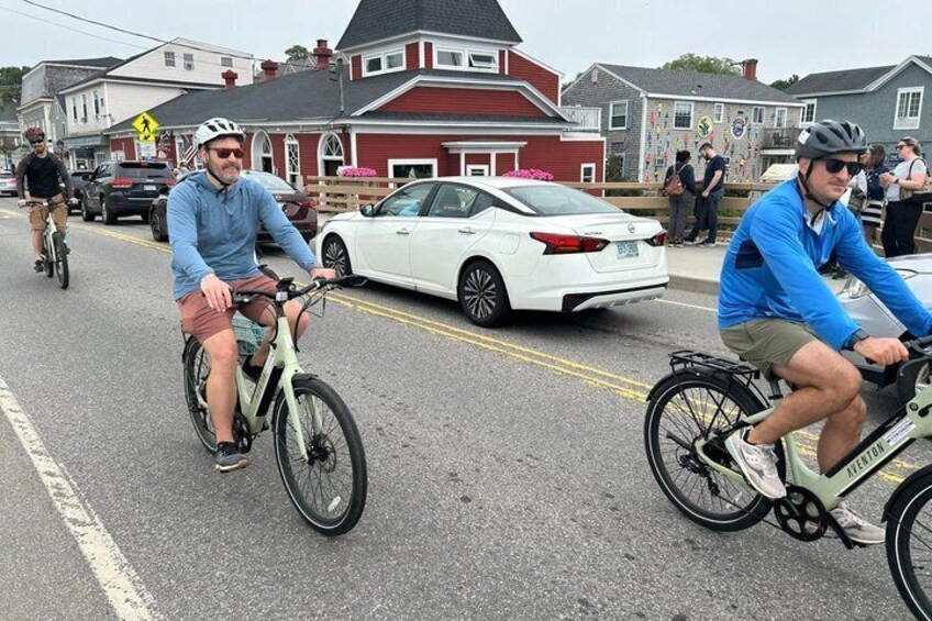 Two cyclists on a road with cars and houses in the background. 5