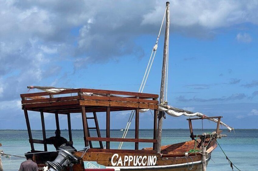 A wooden boat named 'CAPPUCCINO' with a simple structure and a sail, floating on calm waters. 3