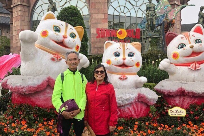 A person in a neon green jacket and another in a red coat standing in front of large cat statues with a building in the background. 4