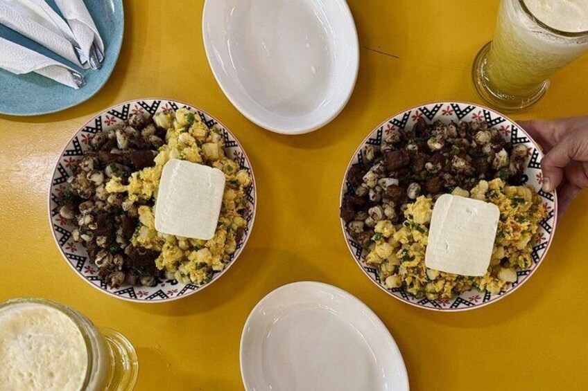 Two plates of food with a block of cheese on top, a glass of a yellow beverage, and a napkin on a yellow table. 1
