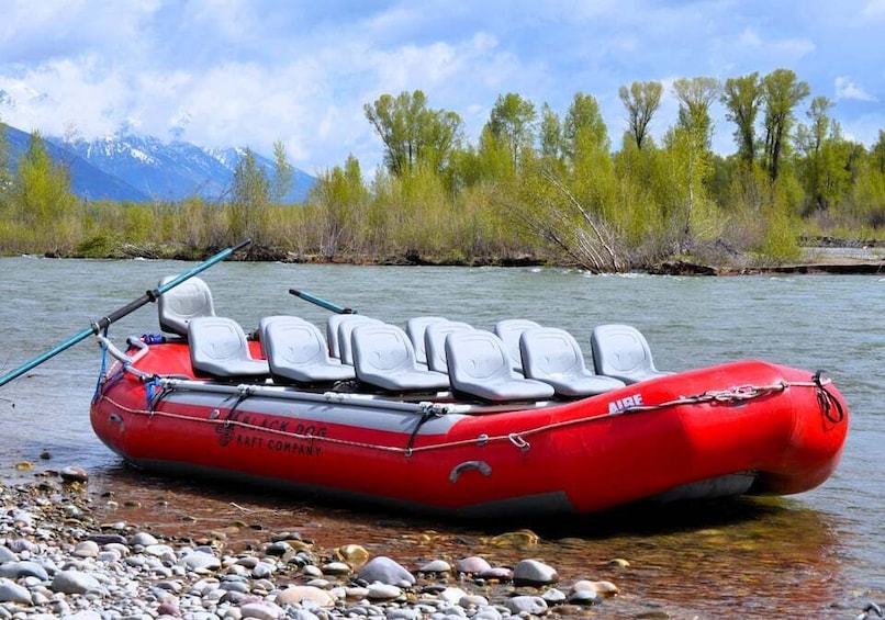 A red inflatable raft with seats and a paddle, docked at a riverbank with rocks and trees in the background. 1