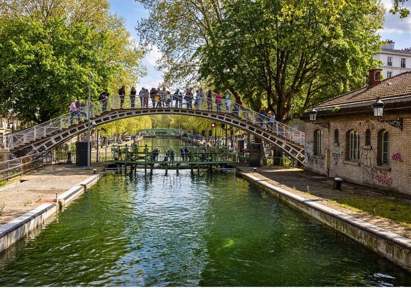 A pedestrian bridge over a canal with people walking, surrounded by trees and buildings. 1